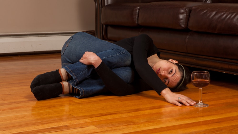 Alcoholism - North Jersey Recovery Center - A woman lays on the floor in front of her couch curled up in a ball as she reaches out for a glass of wine. Her alcohol addiction is out of control and she needs alcoholism rehab.