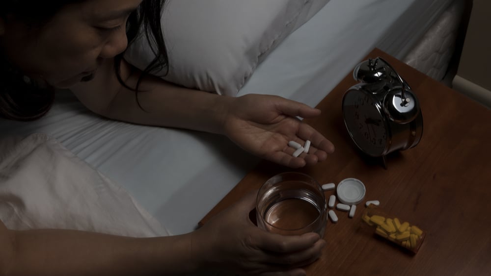 How Long Do Opioids Stay in Your System? North Jersey Recovery Center - A woman is laying in bed and reaching for multiple opioids on her bedside table, which leads her to wonder, "How long to opiates stay in your system?" and if she needs to seek professional treatment.