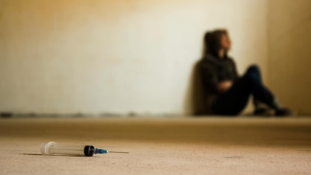 How Much Do Drugs Cost: The Steep Price of Addiction North Jersey Recovery Center - A homeless young male is sitting next to a building on the street with drugs and a syringe near him as he contemplates how much he has spent on drugs based on a street drug prices chart and how he can overcome his addiction and homelessness.