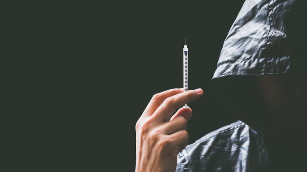 Morphine Addiction and Abuse - North Jersey Recovery Center A man stands to the right side of the image in a dark jacket with the hood pulled up his hand is raised up by his darkened face with a needle of morphine.