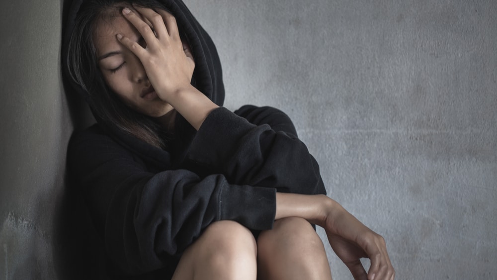 Adderall Withdrawal and Detox North Jersey Recovery Center - A young woman is sitting against a wall with her hood up and palm in her face as she begins to experience Adderall withdrawal symptoms.