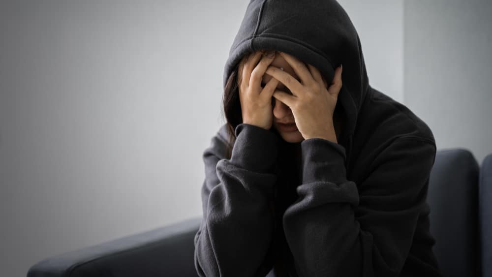 Methadone Withdrawal and Detox North Jersey Recovery Center - A young woman is beginning to feel the effects of methadone withdrawal as she sits on her couch with her hood up and her face in her hands.