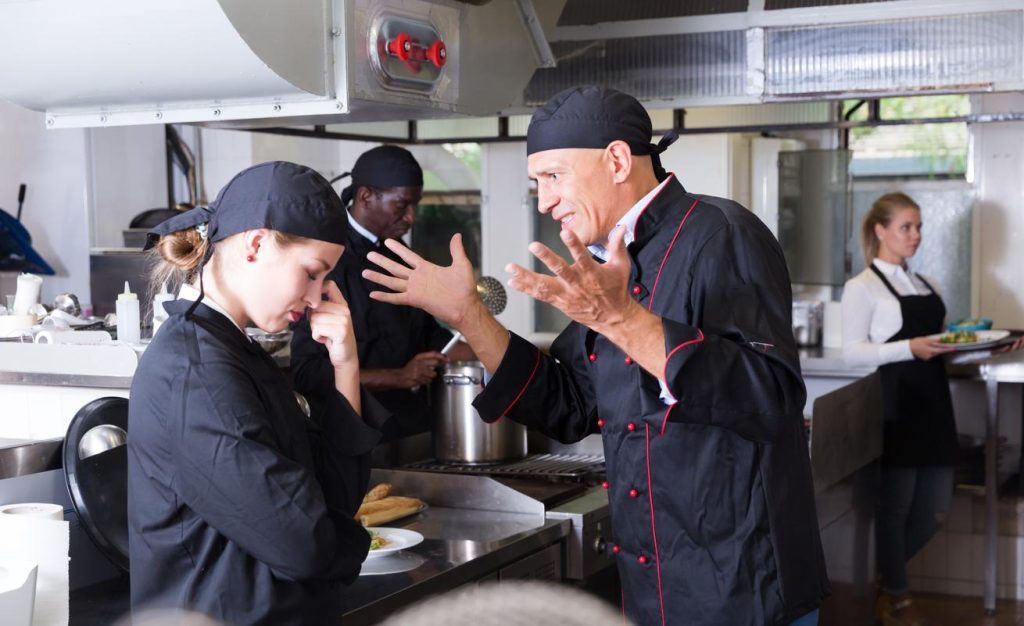 Enraged chef expressing dissatisfaction with work of frustrated girl in restaurant kitchen - drug abuse among chefs